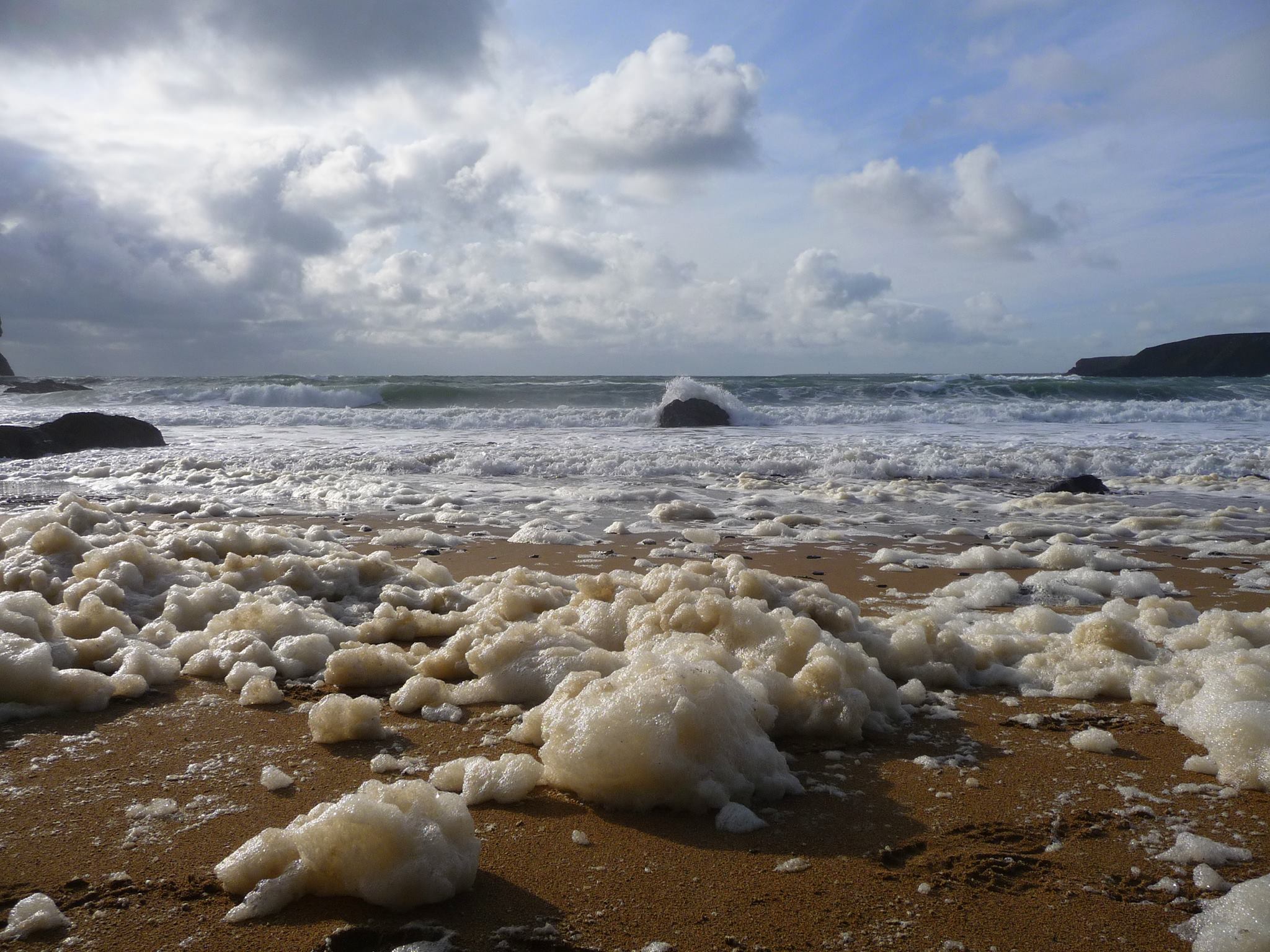 A collection of off white coloured foam on a sandy beach with waves in the background